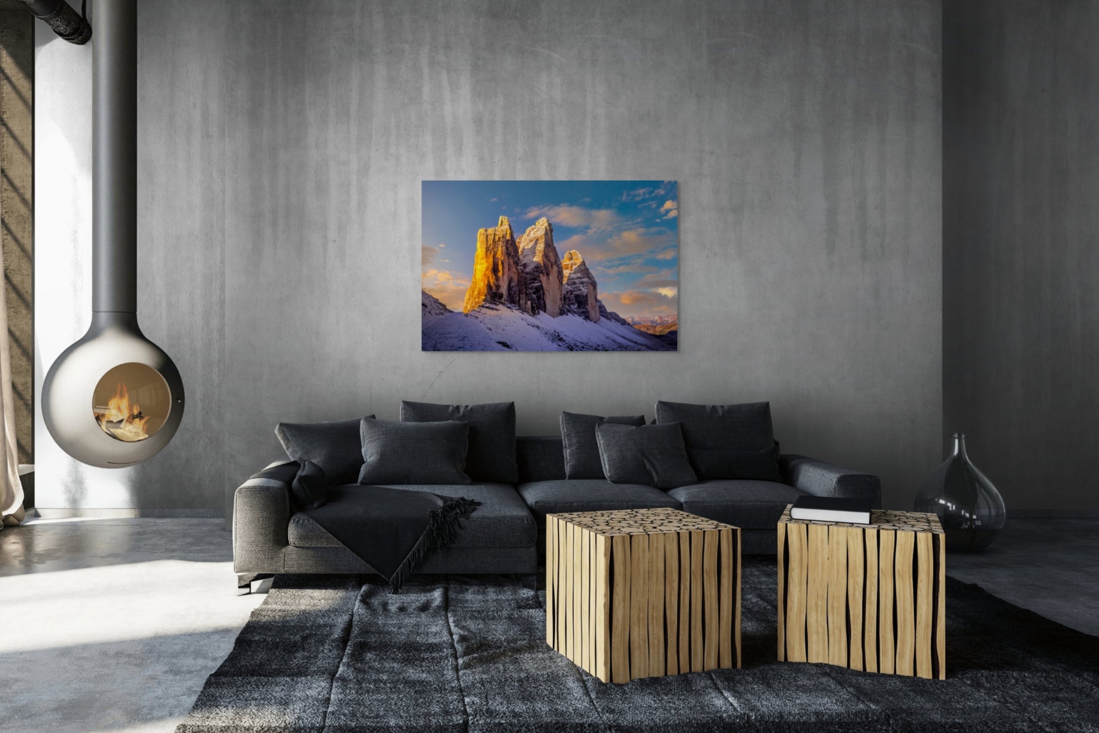Fine art mountain photograph “Cold Fire on Tre Cime di Lavaredo (Drei Zinnen)” by Sherry Keene displayed in a modern luxury living room with concrete walls, gray sofa, and suspended fireplace. The Dolomites peaks glow with golden sunrise after a snowstorm — museum-quality wall art perfect for alpine homes, ski cabins, vacation houses, or collectors seeking large mountain landscape art with warmth and contrast.