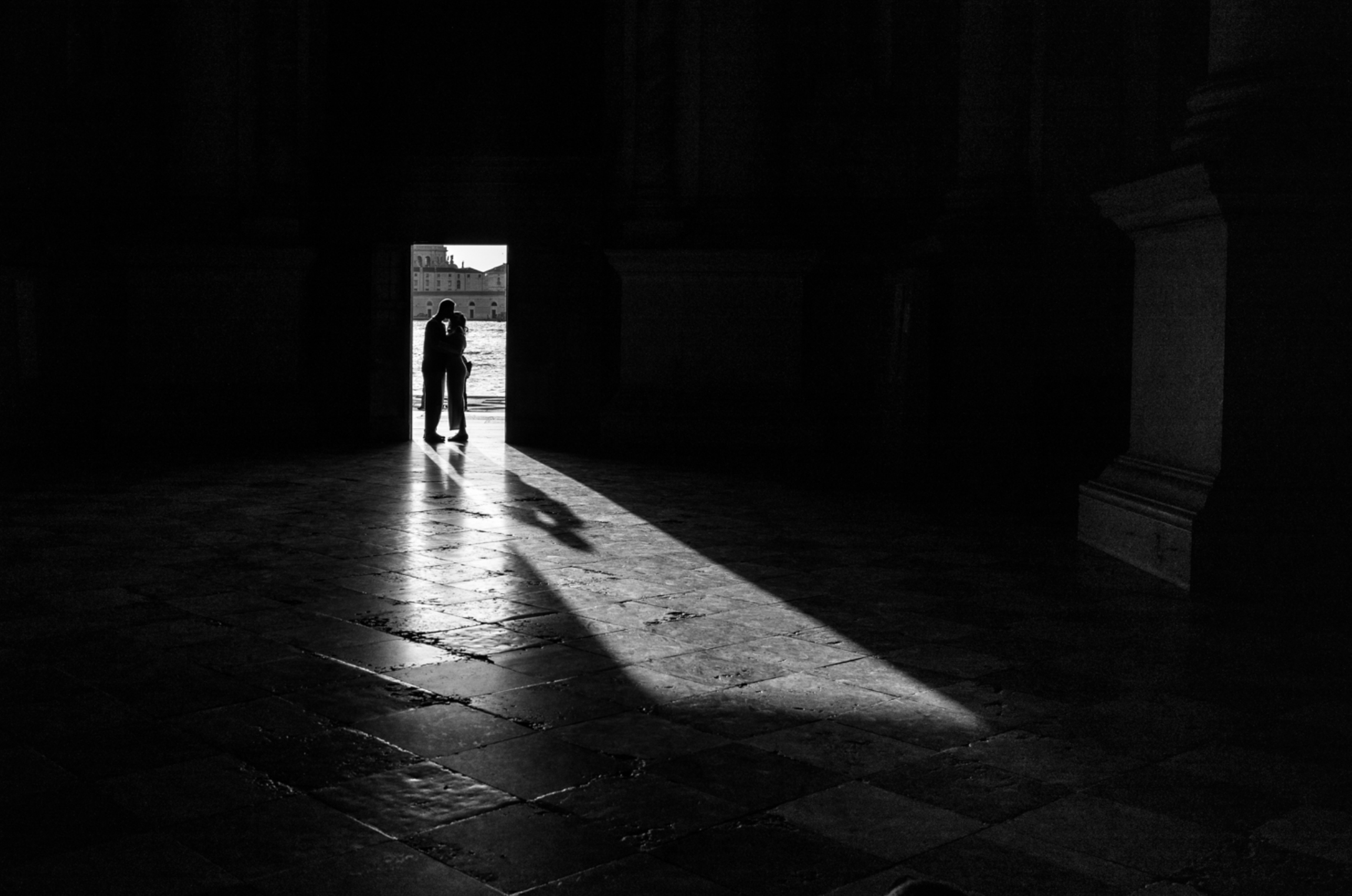 Black and white photograph Two Souls in Sacred Stillness by Sherry Keene – Refocus Award Honorable Mention – two silhouetted figures framed in light beneath the archways of San Giorgio Maggiore, Venice; an emotional, museum-quality fine art print that speaks to connection, stillness, and timeless European beauty